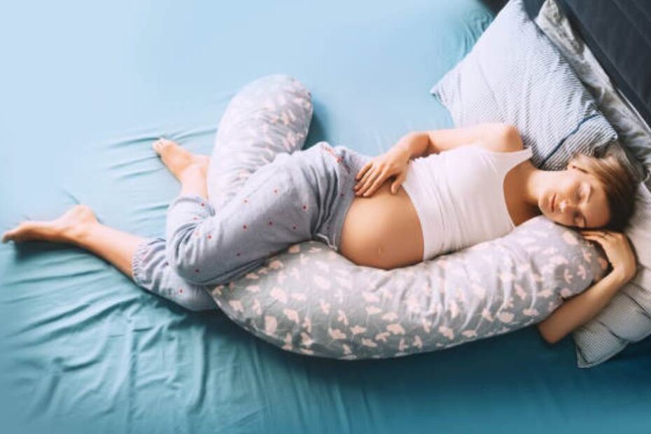 Pregnant woman smiling while hugging a U-shaped full body maternity pillow in a cozy bedroom.